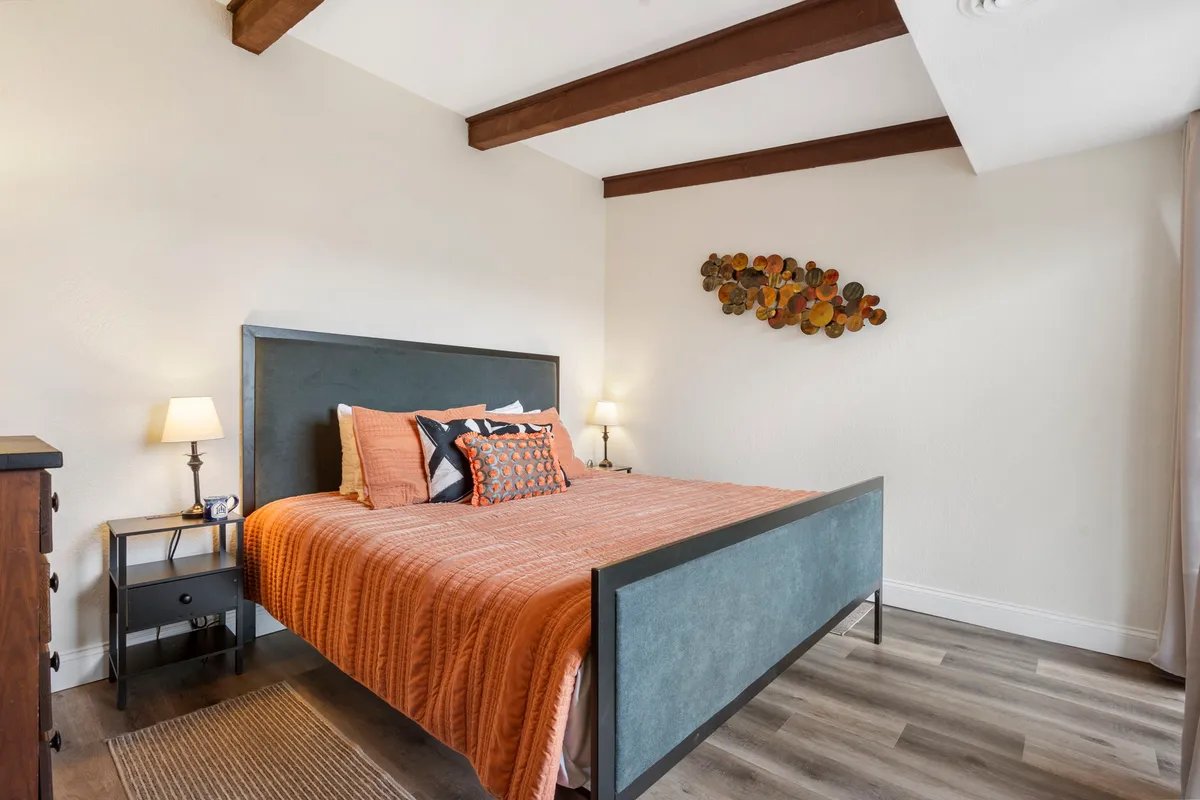 A warm and welcoming bedroom featuring a bed with vibrant orange bedding, decorative wall art, and a nightstand with a lamp. Sunlight filters through the window, creating an inviting atmosphere perfect for rest and relaxation.