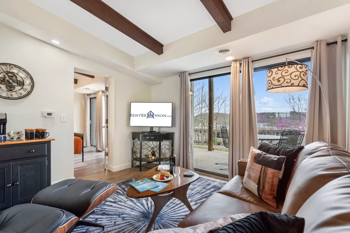 A bright and cozy living room featuring a comfortable sofa, a coffee table with snacks, a large glass door leading to the outside patio, a TV, and a decorative wall clock. Sunlight streams in through the glass door, creating a warm, inviting space ideal for relaxing or gathering with friends and family.