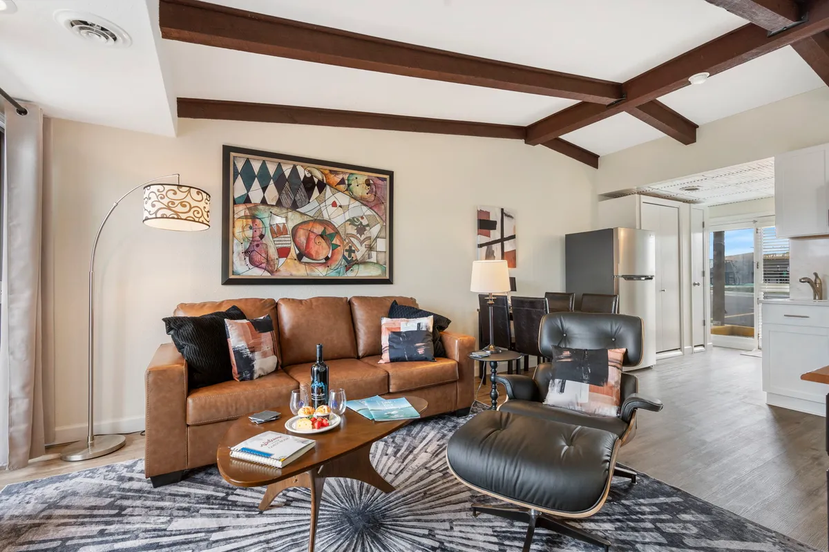 The image showcases a stylish living room with a plush brown leather sofa, vibrant artwork adorning the wall, a sleek black armchair, and a chic coffee table set for sharing wine. The soft lighting and inviting arrangement create an atmosphere perfect for both relaxing and entertaining.