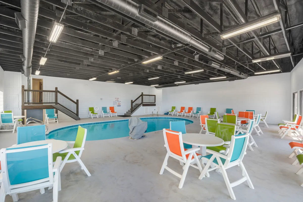 A lively indoor swimming pool area with vibrant, colorful chairs and tables lining the deck. The large windows fill the space with light, and guests are seen swimming and relaxing by the water, creating an inviting year-round leisure atmosphere.