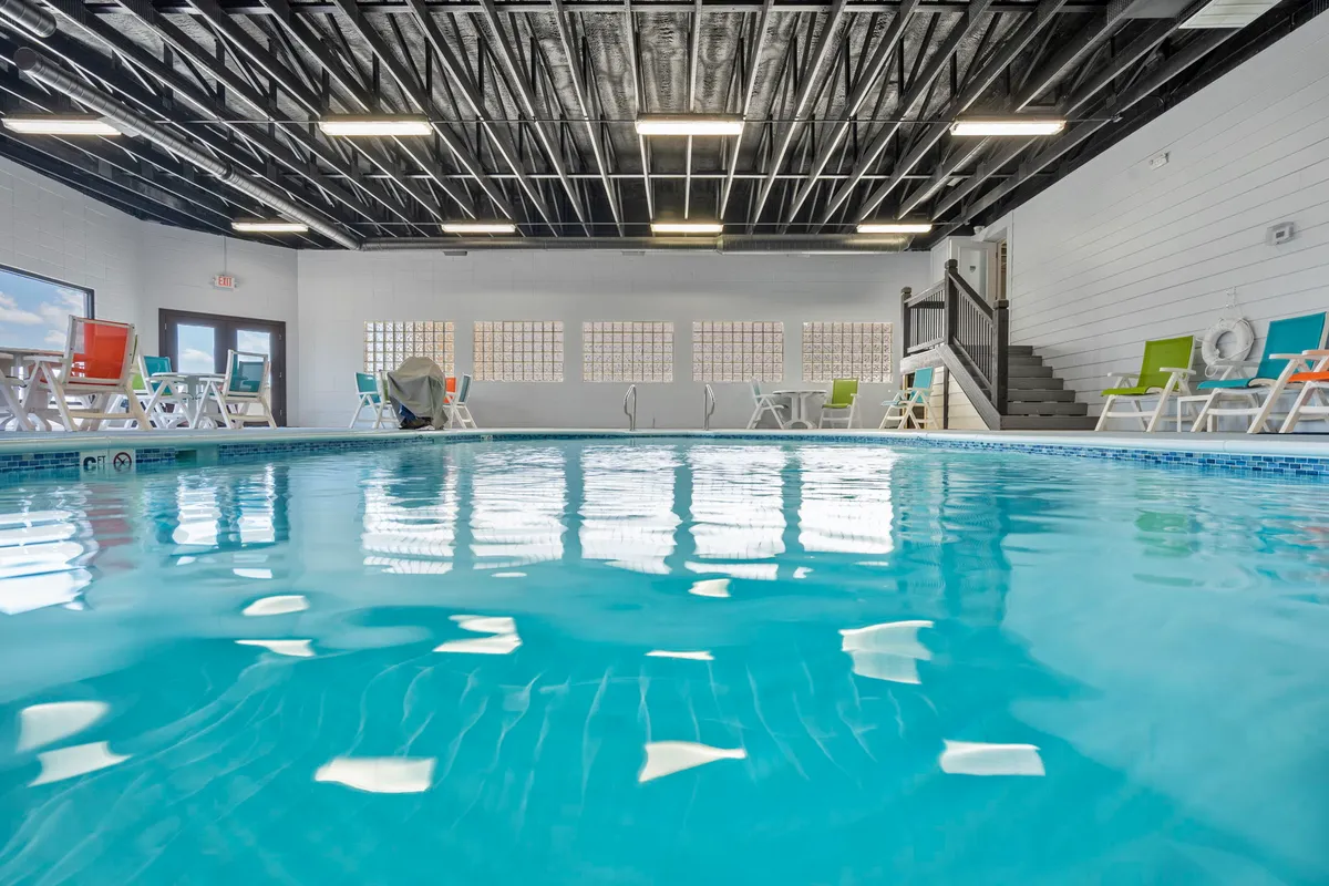 A bright and tranquil indoor pool area with colorful lounge chairs and a staircase leading into the inviting water. Large windows reveal glimpses of the outside greenery, connecting the pool environment to the natural surroundings.