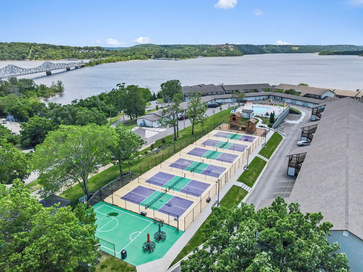 This vibrant exterior shot captures a lively scene with tennis courts, a sparkling swimming pool filled with families, views of the lake in the background, and a playground where children are having fun. The setting is illuminated by the warm glow of sunset, adding an atmosphere of joy and community.