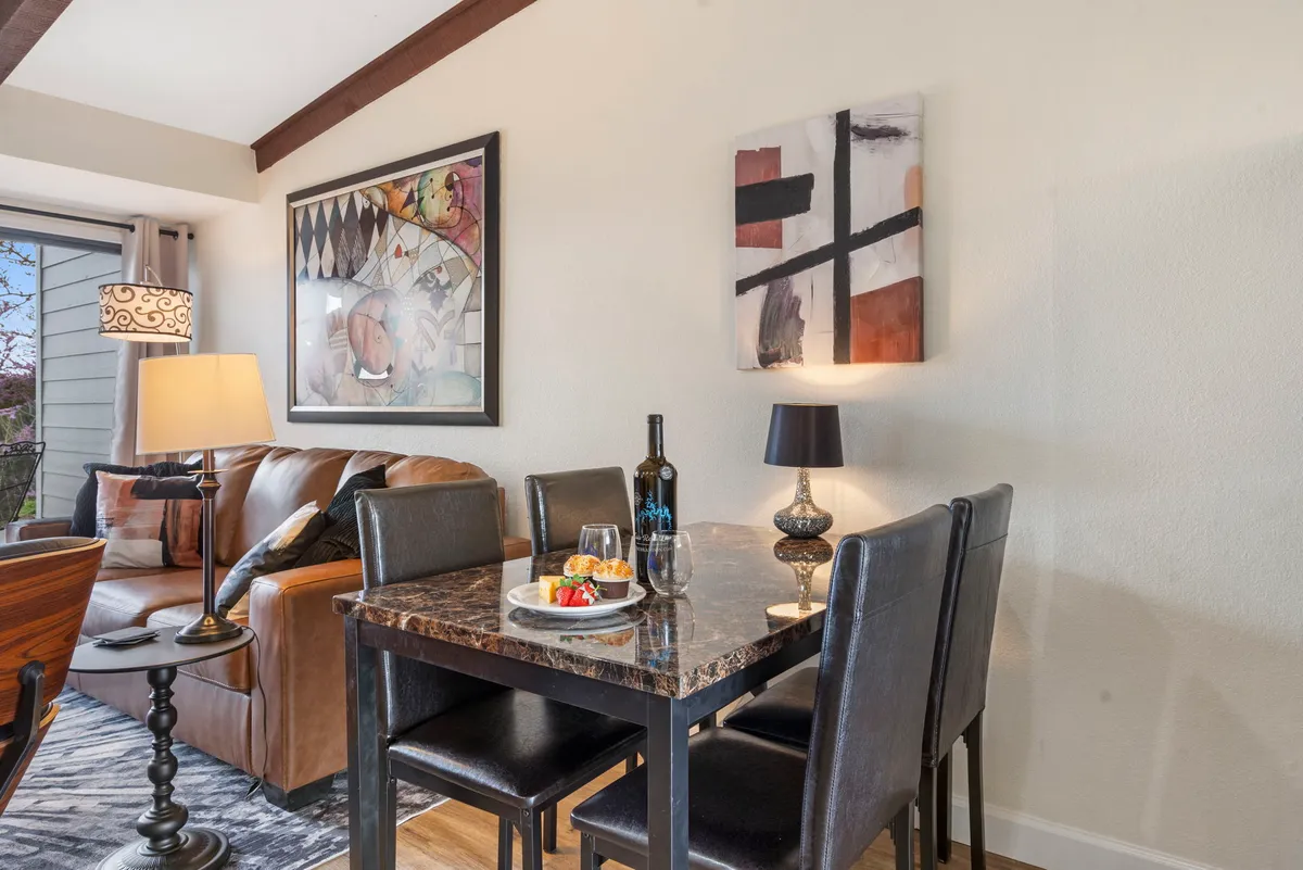 A welcoming dining area highlighted by a table set with food and wine, comfortable seating, artwork on the wall, and a sofa with soft cushions in the background. The scene evokes warmth and connection, ideal for sharing meals and conversation.