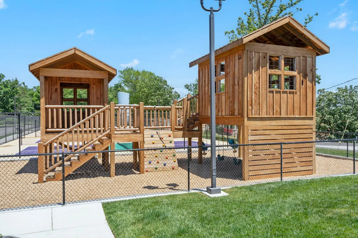 A vibrant playground scene outdoors, featuring wooden playhouses, a climbing wall, and a swing set, with children energetically playing and socializing in a sunny, safe environment. The mood is joyful and active, ideal for families with kids.