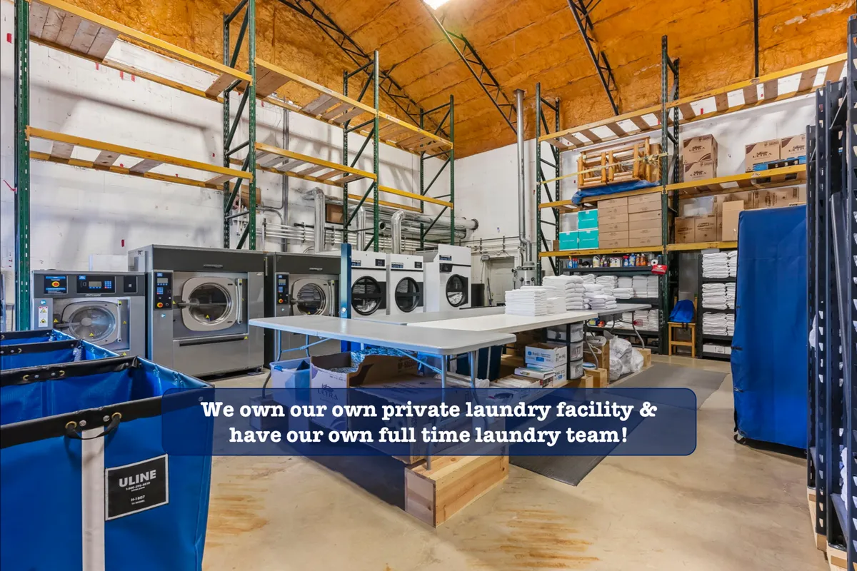 A bright and organized interior view of industrial washing machines, laundry baskets, and clean storage shelves. The scene conveys efficiency and professionalism in laundry care, suggesting a high standard for cleanliness and guest comfort.