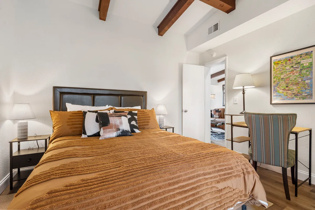 A stylish bedroom with textured bedding and decorative pillows, complemented by side tables with lamps. There is a desk and chair in a striped design and a framed map on the wall, suggesting both comfort and functionality.