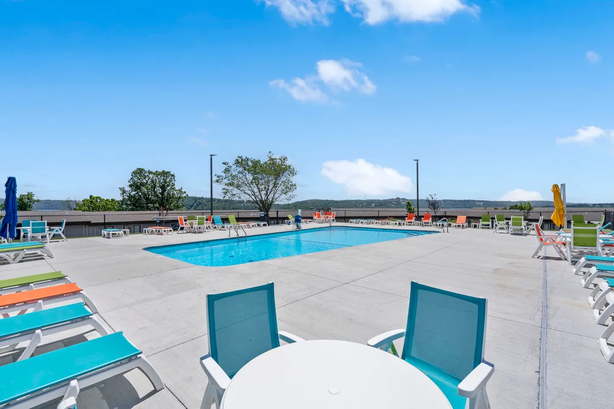 An outdoor swimming pool surrounded by colorful lounge chairs and picturesque landscaping. People are seen sunbathing, swimming, and socializing, capturing the leisurely pace of summer days.