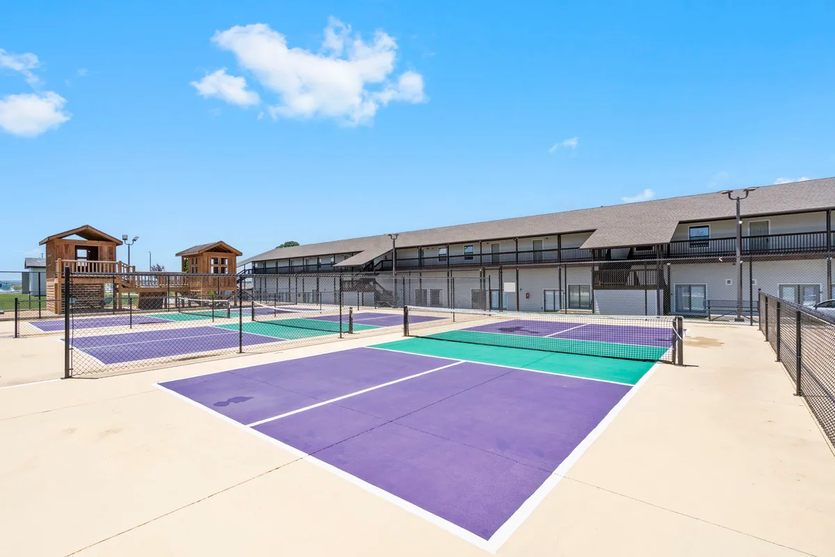 A spacious exterior view highlighting tennis courts (adaptable as pickleball), a vibrant playground structure, and a multi-unit condo building in the background. Children are playing, and families gather, illustrating active recreation and community.